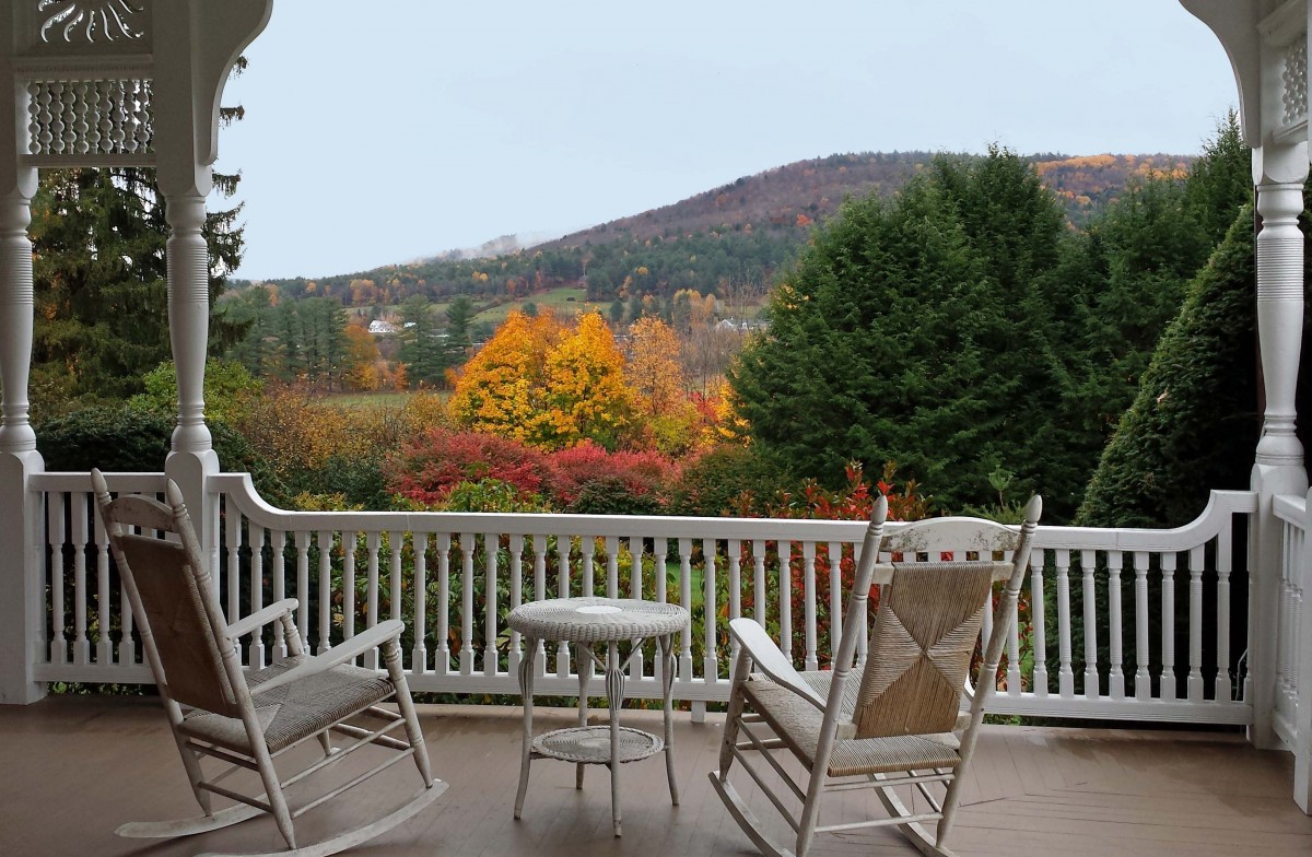 Two white wooden rocking chairs sit on a matching balcony overlooking colorful autumn trees and small forested hills.