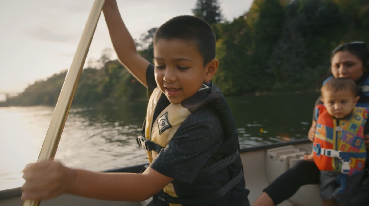 Young boy paddles boat through water with family in back.