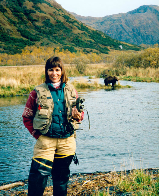 Mollie Beatie - a woman with brown hair wearing fishing gear - stands on a riverbank holding binoculars as a bear stands in the background.