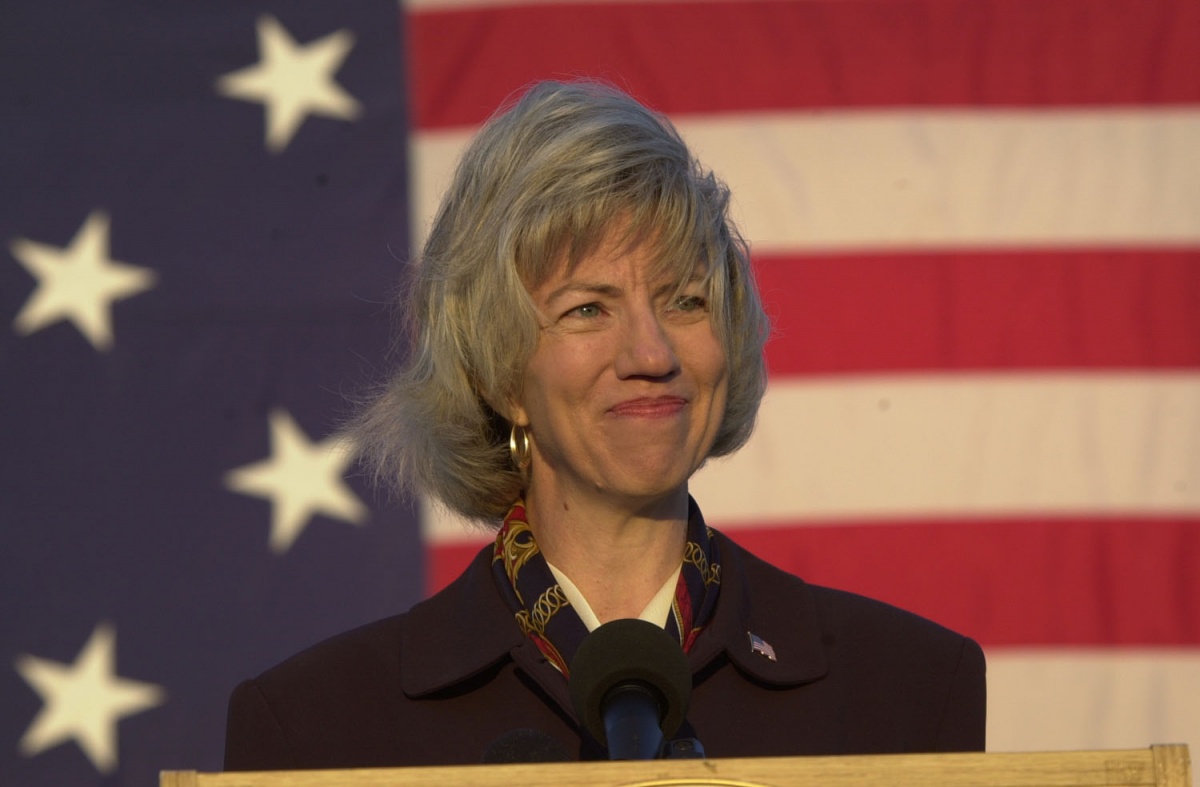 Gale Norton - a woman with blond hair wearing a scarf and coat - stands in front of the American flag.