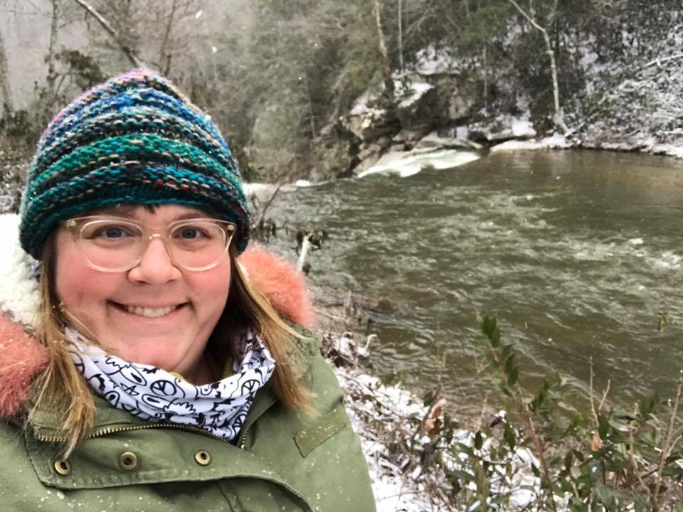 Elizabeth Smith, a blond white woman wearing a heavy coat, glasses and a knit cap, stands in front of a river on a snowy day. 