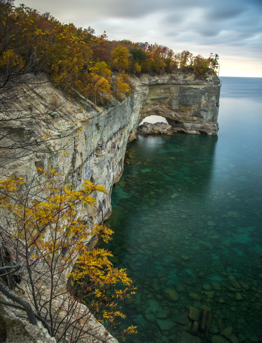 Trees grow on top of a stone cliff rising above the blue waters of a large lake.