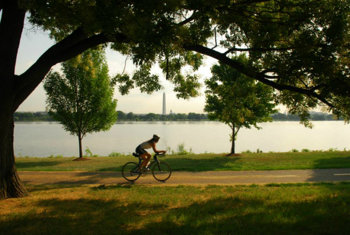 A biker rides along a paved path with a river and the Washington Monument in the background