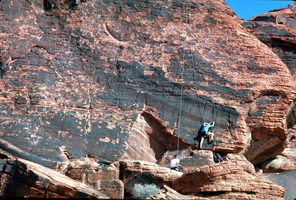 Two people with climbing gear and ropes climb up a large red rock cliff.