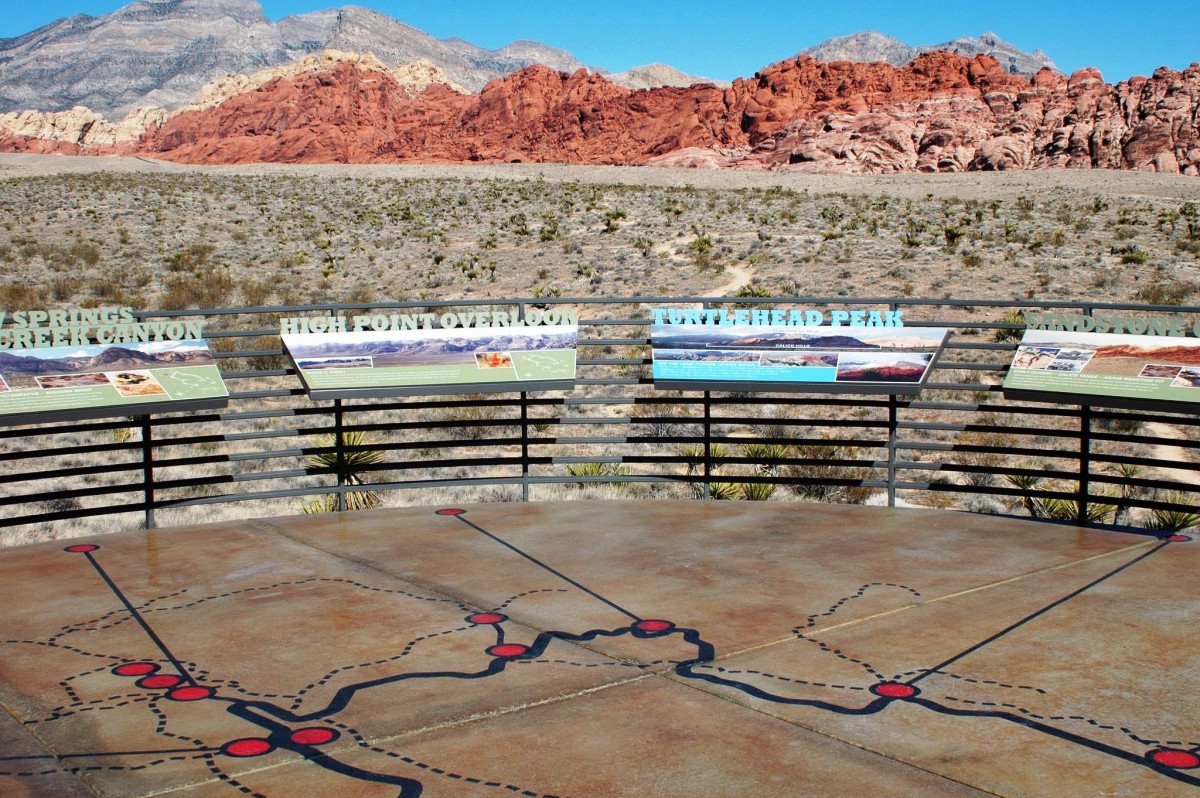 A concrete platform with a map drawn on it is surrounded by a low fence holding up several information plaques describing the views in the distance of red rock mountains.