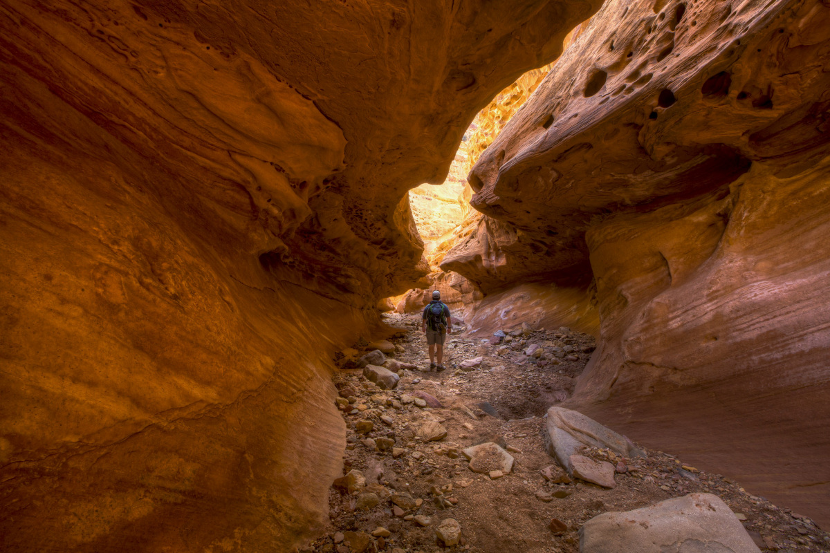 A man with a backpack hikes through a narrow red rock canyon.