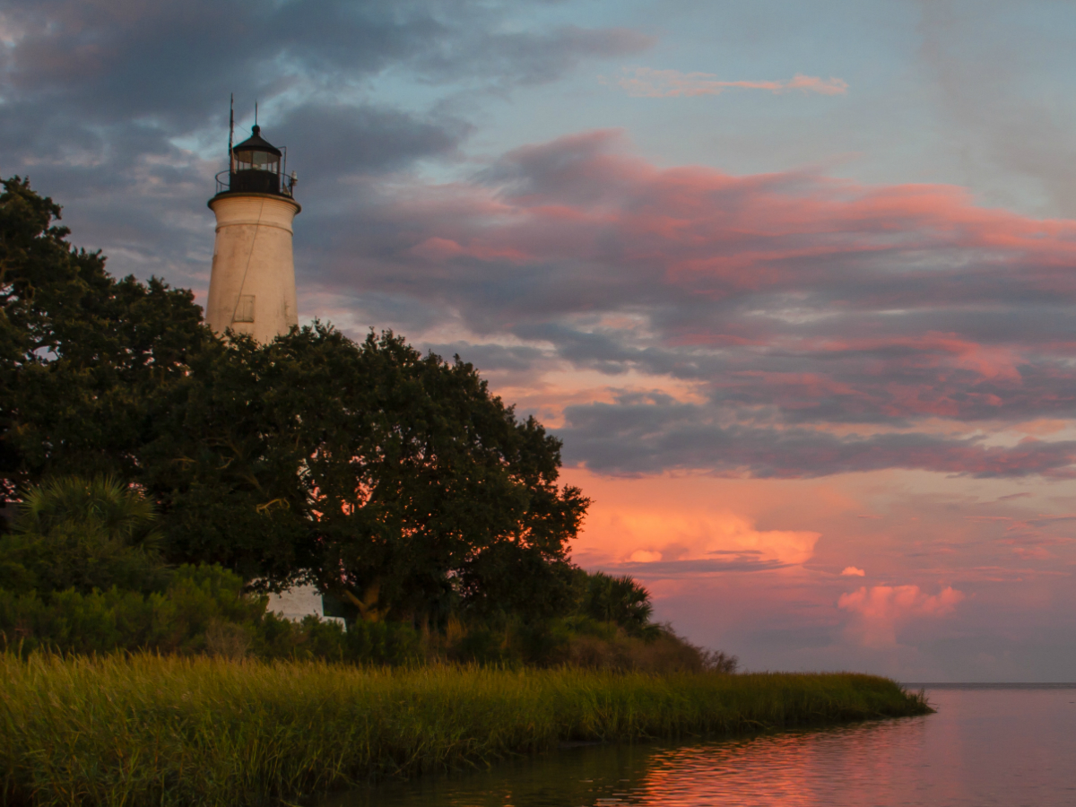 A white lighthouse rises above some trees on the grassy bank of a bay under a sunset sky.