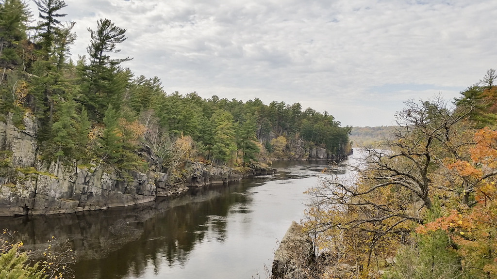 A calm, narrow river flows past a short, rocky cliff with trees on top.