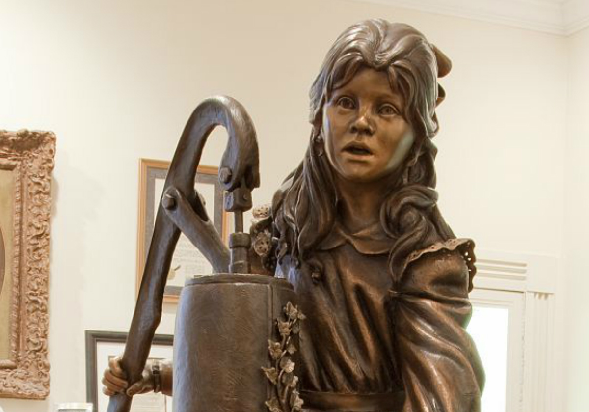 A bronze statue of a little girl in a dress standing next to a water pump.