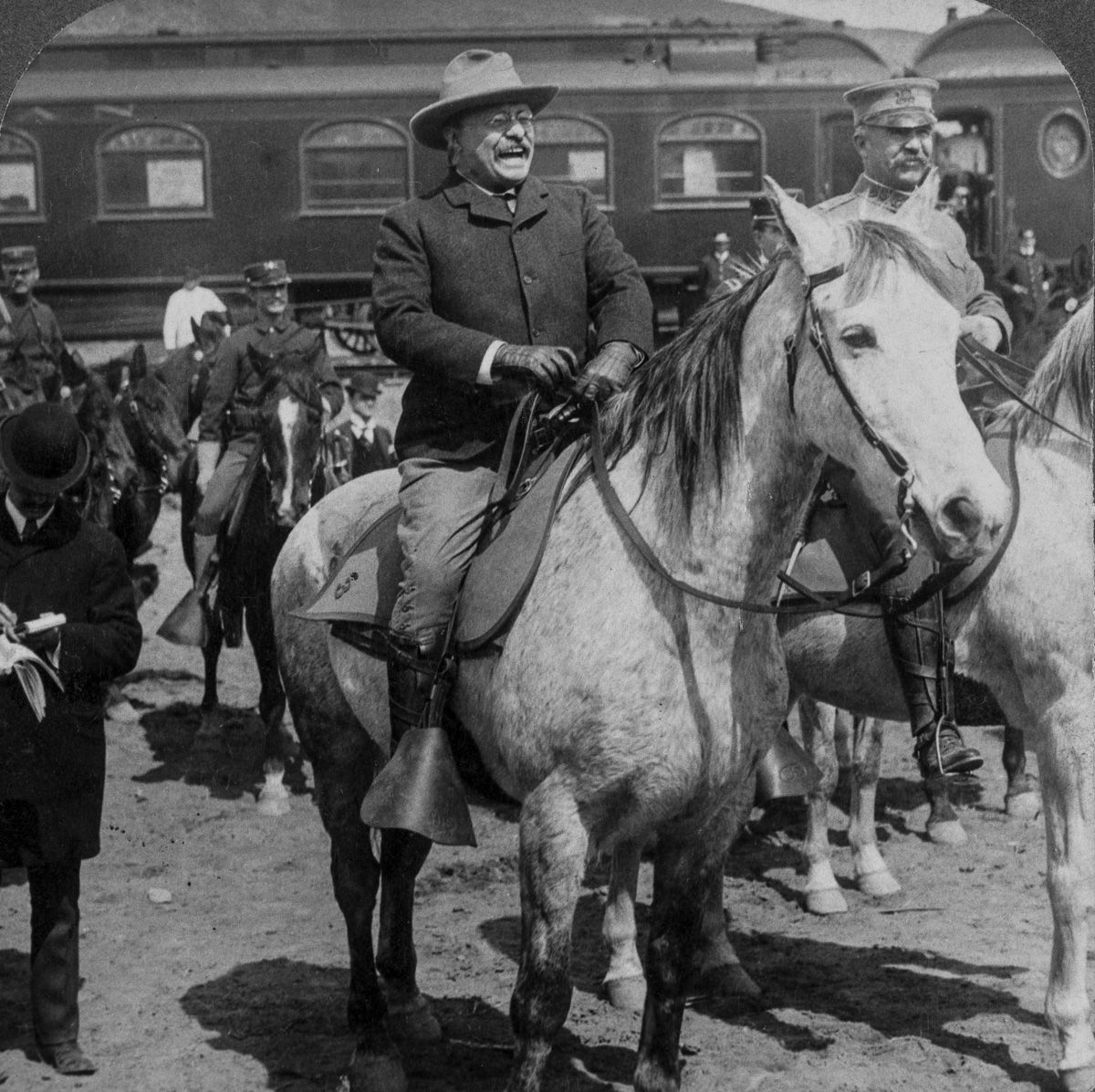 Grainy photo of Theodore Roosevelt riding a horse surrounded by a crowd at Yellowstone National Park.