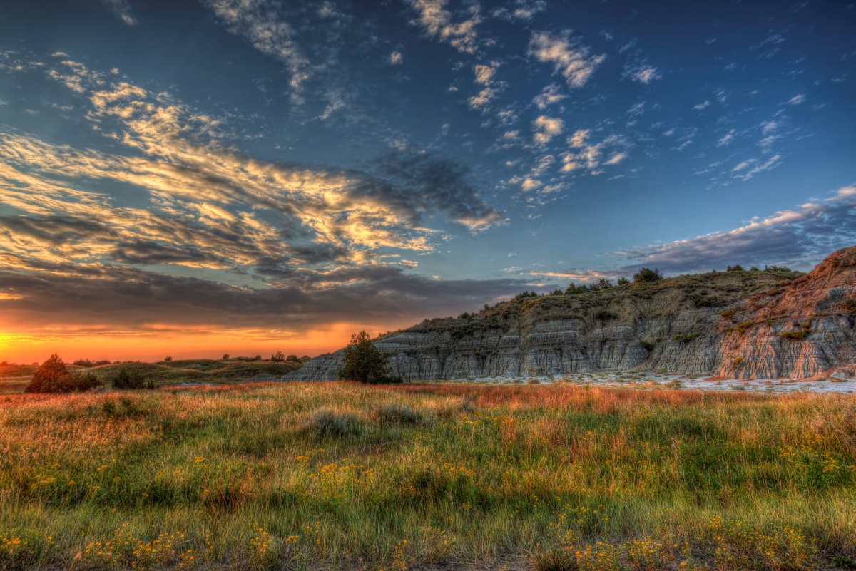 The sun sets over a green grassy plain with a colorful bluff rising in the background.