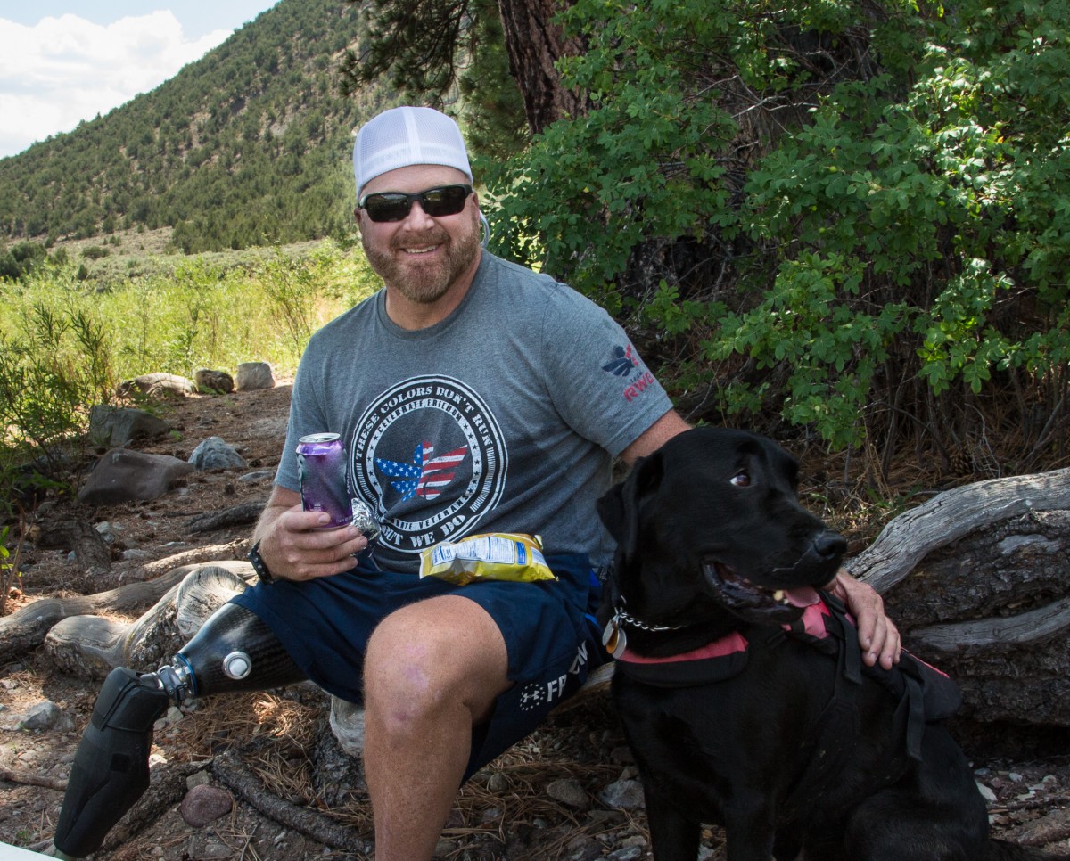 A man sits and snacks in the shade next to his Labrador Retriever on a fallen log.