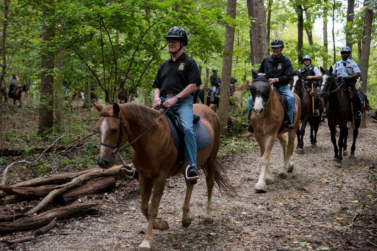 Veterans mounted on horses proudly walk in a line with Park Police on a path winding through tall green trees.