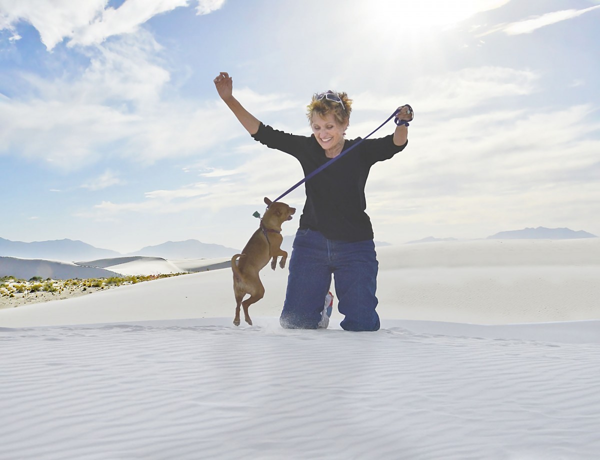 A woman with short blond hair smiles as her small brown dog jumps around her on top of a wide white sand dune.