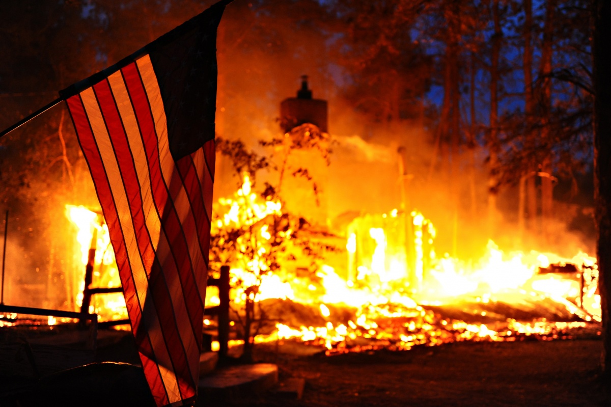 An American flag hands on a pole in front of a burning house.